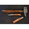 Un martillo rústico con mango de madera desgastada y cabeza de metal se encuentra junto a dos navajas plegables Opinel, entre ellas un Cuchillo Opinel N°7 VRN de Acero Carbono (uno abierto y otro cerrado) sobre una superficie oscura y texturizada.