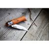Un cuchillo Opinel N°8 VRN de acero carbono, con mango de madera y hoja de metal brillante, reposa sobre una superficie de madera desgastada. Cerca, una rama de árbol seca y delgada realza la escena rústica y el mango muestra de forma destacada el grabado "Opinel".
