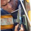 Una persona está profundamente concentrada en aflojar un accesorio de metal unido a un componente de madera y metal de un barco utilizando el Cuchillo Opinel N°8 Outdoor, que cuenta con una hoja dentada y un mango con detalles en azul y negro.