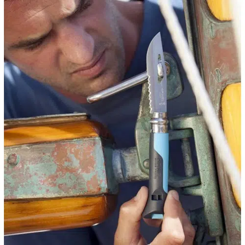 Una persona está profundamente concentrada en aflojar un accesorio de metal unido a un componente de madera y metal de un barco utilizando el Cuchillo Opinel N°8 Outdoor, que cuenta con una hoja dentada y un mango con detalles en azul y negro.