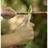 Un par de manos tallan hábilmente una pequeña rama con un cuchillo Cuchillo Opinel N°8 Outdoor sobre un fondo de vegetación borrosa, lo que sugiere un entorno al aire libre o de carpintería.
