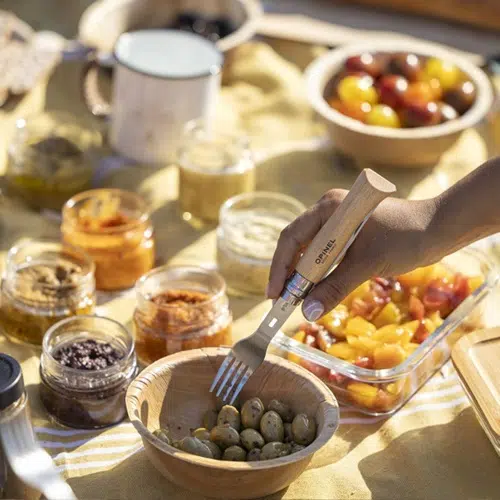 Una mano usa un tenedor para recoger aceitunas de un cuenco de madera en un encantador entorno para picnic. Alrededor del cuenco hay frascos de crema para untar, un recipiente de vidrio con tomates cherry de colores y el Set Completo Opinel para Picnic de 4 piezas presentado sobre un mantel amarillo brillante.