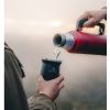 Un termo Mate Stanley Classic de 236 ml en color rojo vierte líquido caliente en una taza negra sostenida por una persona vestida con una chaqueta marrón, frente a un fondo exterior con niebla.