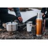 Junto a un río tranquilo, dos personas se sientan en las rocas mientras su juego de preparación y cocción Stanley Adventure Prep + Cook Set de 1,5 l cocina a fuego lento una comida en una estufa de camping. Dos tazas de marca y un termo completan la escena, prometiendo una experiencia gastronómica perfecta al aire libre.