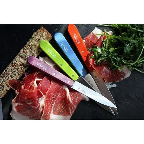 Un colorido juego de cocina de 4 piezas Opinel N°112 Office brilla sobre una superficie de pizarra, rodeado de finas lonchas de carne curada, ensalada verde fresca y un trozo de pan con semillas. El juego incluye cuchillos con mangos vibrantes en azul, naranja, verde y violeta, cada uno de los cuales muestra de forma destacada la marca "Opinel".