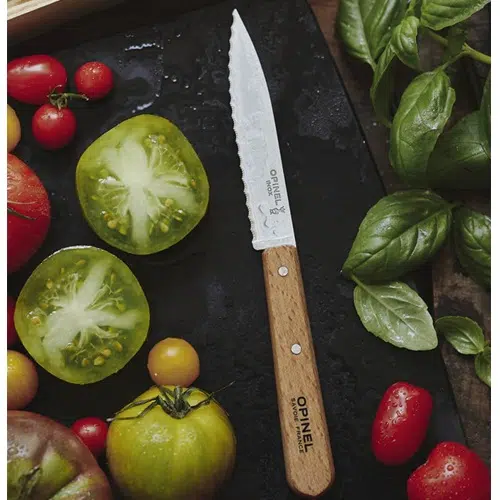 Un Cuchillo Opinel N°113 Dentado reposa sobre una tabla de cortar negra, acompañado de tomates frescos y coloridos y hojas de albahaca. Algunos de los tomates han sido cortados en rodajas para resaltar sus semillas y su interior brillante.