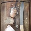 Una tabla de cortar de madera con un cuchillo dentado Opinel N°116 de Pan Parallèle, una barra de pan redonda entera y dos rebanadas. Se encuentran esparcidas semillas de chía y de girasol, que complementan el encanto rústico de la escena. El pan luce una ligera capa de harina por encima.