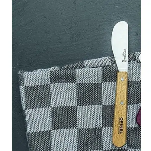 Un cuchillo Opinel N°117 Mantequilla, con mango de madera, se encuentra elegantemente colocado sobre una tela a cuadros blancos y negros. Un texto grabado adorna la hoja del cuchillo, que reposa sobre lo que parece ser una superficie oscura y texturizada que podría ser pizarra.