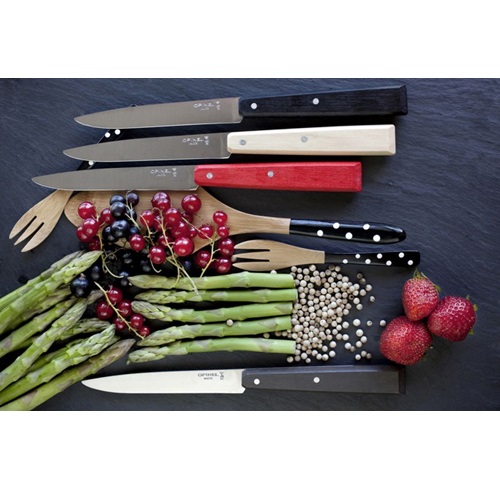 El juego de 4 cuchillos Opinel N°125 Bon Apettit Loft, con sus característicos mangos de madera, se presenta con buen gusto junto a una variedad de utensilios, espárragos frescos, manojos de grosellas, fresas y granos de pimienta blanca sobre un fondo oscuro.