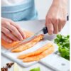 Una persona corta un filete de salmón fresco con el afilado Cuchillo Opinel N°221 Afilado Intempora sobre una tabla de cortar blanca. Cerca se ven parcialmente cilantro fresco y otros ingredientes, lo que sugiere que se está preparando una comida.