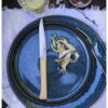 Una mesa con un plato de cerámica azul elegantemente acompañado de un cuchillo y un tenedor con mango de madera de la colección de 4 piezas Fracette Mango de Fresno de Caja de Cuchillos Opinel. En el plato se encuentran artísticamente dispuestos champiñones en rodajas y ralladura de limón, mientras que en el fondo, un vaso de agua de lima reposa sobre un mantel blanco rústico.