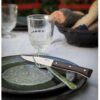 Una mesa con un plato verde con un cuchillo y un tenedor, probablemente del elegante juego de 4 piezas Fracette Mango de Fresno Oscuro de Caja de Cuchillos Opinel, colocado encima. Cerca hay un vaso transparente y al fondo hay una cesta llena de pan. El mantel es de color claro.