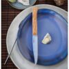 Sobre un plato azul reposa una Caja de Cuchillos Opinel Mango de Olivo Fracette 4 piezas, acompañada de un trocito de queso. En el fondo, un tenedor y otro plato reposan sobre la superficie de madera, completando esta rústica disposición de la mesa.