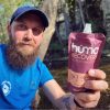 Un hombre con barba, con camisa azul y sombrero, sostiene una bolsa de Gel Energético Hüma Recovery Smoothies Mixed Berries. Está al aire libre, rodeado de naturaleza, con árboles y la luz del sol filtrándose. La atención se centra en el producto que tiene en la mano.