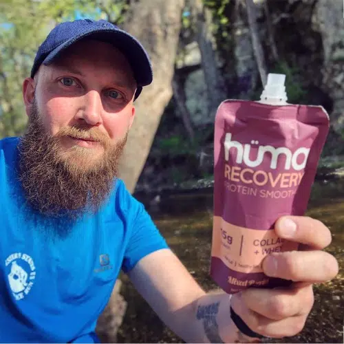 Un hombre con barba, con camisa azul y sombrero, sostiene una bolsa de Gel Energético Hüma Recovery Smoothies Mixed Berries. Está al aire libre, rodeado de naturaleza, con árboles y la luz del sol filtrándose. La atención se centra en el producto que tiene en la mano.