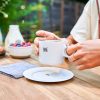 Una persona sostiene una taza Stanley Cappuccino Daybreak con Plato de 192 ml, adornada con un pequeño logotipo de dragón, sobre un platillo blanco en una mesa de madera. Cerca hay un bol con bayas, una botella de leche y una servilleta azul. El fondo presenta una vegetación borrosa que realza el encanto matutino de la hora del capuchino.