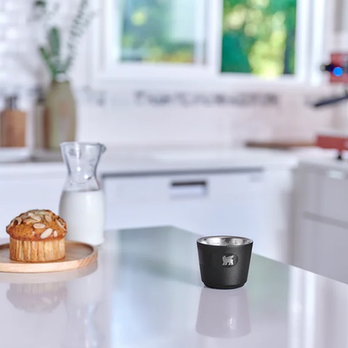 En una cocina contemporánea, una taza Stanley Espresso Daybreak de 65 ml ocupa un lugar destacado sobre la encimera blanca. Detrás de ella, se puede ver una jarra de leche y un panecillo sobre un plato de madera. La luz natural entra a raudales por la ventana, lo que realza el ambiente acogedor del espacio.