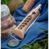 El cuchillo Opinel N°10 con abrebotellas y sacacorchos está colocado sobre un paño azul, rodeado de rebanadas de pan, salchichas y queso blanco. También se ve un corcho, lo que sugiere un picnic al aire libre sobre el césped.