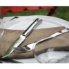 La mesa presenta un tenedor y un cuchillo del juego de 4 piezas de la colección Perpétue de Opinel, colocados elegantemente sobre una servilleta marrón encima de un plato blanco. Una silla roja se asoma a la vista contra un fondo de césped exuberante, evocando el encanto de un comedor al aire libre.