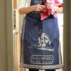 En la cocina, una persona con un delantal azul que recuerda al modelo Delantal Opinel Adulto, sostiene un paño rosa y destila elegancia y profesionalidad.