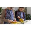 Dos niños con delantales azules Opinel Adulto de Delantal con círculos blancos están de pie frente a una encimera de cocina, cada uno pelando y cortando fruta con cuidado con un cuchillo Opinel, concentrados en sus tareas. El fondo presenta elementos de cocina y vegetación, creando un ambiente culinario sereno.