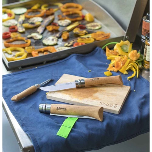 El Kit de Cocina Opinel Nomad, que incluye un cuchillo cerrado y otro abierto, reposa sobre una tabla de cortar de madera encima de un paño azul. Hay cáscaras de verduras esparcidas cerca y verduras cortadas chisporroteando en la barbacoa de fondo.