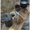 Un ambiente rústico al aire libre cuenta con una tabla de madera que sostiene una cafetera moka, una cafetera negra con líquido, dos tazas, un Cuchillo Opinel N°6 Mango de Olivo y dos rebanadas de pan tostado con mantequilla oscura. El suelo está salpicado de hojas y rocas, lo que realza el ambiente natural.
