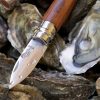 Un Cuchillo Opinel N°9 Ostras con mango de madera y hoja de metal reposa sobre una pila de ostras. La hoja brilla con gotas de agua, mientras que una de las ostras está abierta, revelando su tentador interior.