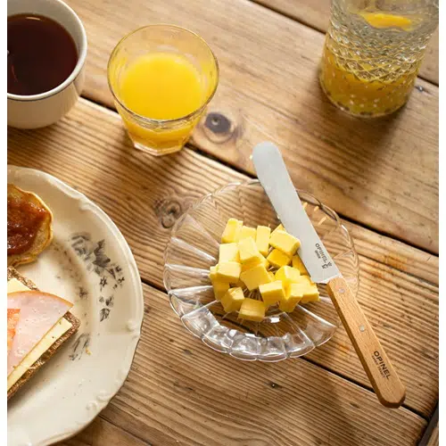 Una mesa rústica de madera preparada para el desayuno muestra un plato con sándwiches abiertos, un vaso de jugo de naranja y una taza de café. Junto a ellos hay un plato de vidrio que contiene mantequilla cortada en cubitos junto con el Cuchillo Opinel para Untar, perfecto para untar.