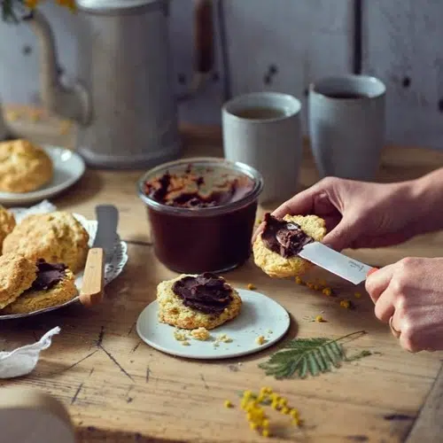Con un cuchillo para untar Opinel, una persona extiende crema de chocolate sobre una galleta. Sobre una mesa de madera se colocan un tarro de chocolate, galletas, dos tazas y una cafetera antigua. Pequeñas flores amarillas y una hoja realzan la acogedora escena con su decoración.