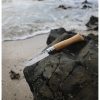 Un Cuchillo Opinel N°9 Mango de Roble está colocado sobre una roca oscura a la orilla del mar, mientras las olas rompen suavemente en la playa de arena al fondo.
