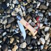 Un Cuchillo Opinel N°8 Emblema de Saboya, adornado con un mango de madera y un cordón rojo, descansa abierto sobre un lecho de piedras lisas y multicolores. El mango está adornado con símbolos grabados y un pequeño Emblema de Saboya, lo que infunde un sentido de tradición y arte a esta elegante pieza.