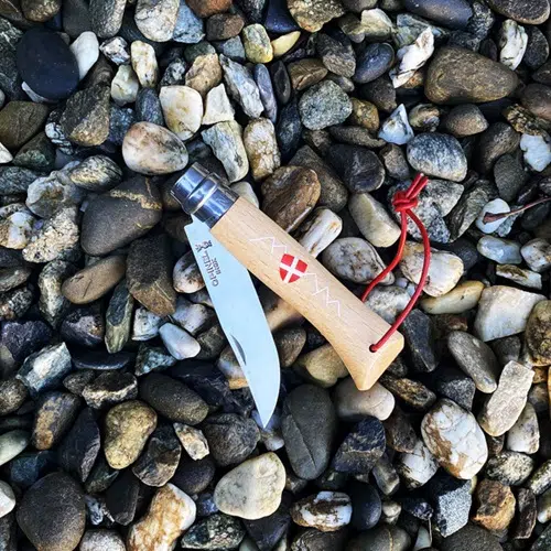 Un Cuchillo Opinel N°8 Emblema de Saboya, adornado con un mango de madera y un cordón rojo, descansa abierto sobre un lecho de piedras lisas y multicolores. El mango está adornado con símbolos grabados y un pequeño Emblema de Saboya, lo que infunde un sentido de tradición y arte a esta elegante pieza.