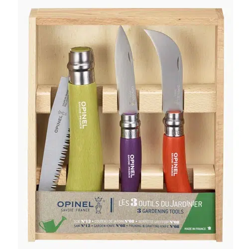 Una caja de madera con la etiqueta "Set Caja Opinel para Jardín" contiene un juego de tres herramientas de jardinería Opinel: una sierra plegable con mango verde, un cuchillo de jardinería con mango morado y un cuchillo de poda con mango naranja. La caja muestra la etiqueta de Opinel en la parte inferior junto con los detalles del producto.