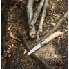 Una Sierra Opinel N°12, con su mango de madera, se encuentra en el suelo junto a algunas ramas cortadas y virutas de madera en un entorno al aire libre. El entorno está cubierto de hojas caídas y virutas de madera, creando un ambiente rústico que refleja un sentido de artesanía en medio de la serenidad de la naturaleza.