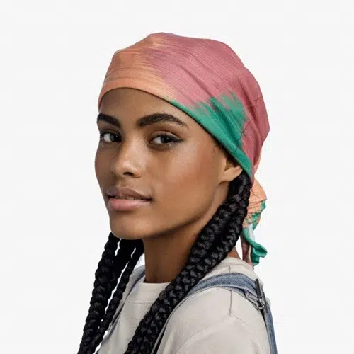 Una persona con largas trenzas mira por encima del hombro, luciendo la vibrante Bandana Buff CoolNet UV en Dilm Multi. Combinada con una camisa y un mono de color claro sobre un fondo blanco liso, su estilo rezuma estilo informal.