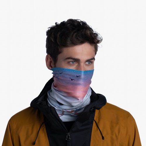 Una persona con cabello rizado lleva una chaqueta naranja y una mascarilla Bandana Buff ThermoNet White Ocean que muestra una escena de playa oceánica blanca con pájaros, sobre un fondo blanco liso.