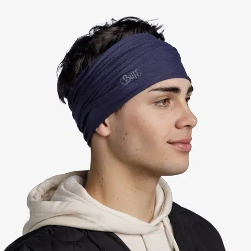 Una persona que lleva una banda para el pelo Bandana Buff Original EcoStretch Solid de color azul oscuro mira hacia la derecha. Tiene el pelo oscuro y lleva una sudadera blanca con capucha y una chaqueta negra sobre un fondo blanco liso.