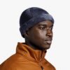 Una persona que viste un gorro Buff Tricot Ervin en azul y una chaqueta naranja con cremallera mira hacia un lado sobre un fondo blanco liso.