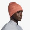 Una persona vistiendo un Gorro Buff Tricot Norval color coral y una chaqueta gris claro, colocada a la derecha contra un fondo blanco sin adornos.