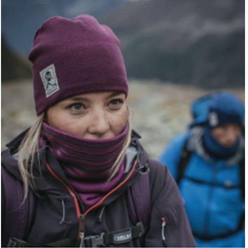 Una persona que lleva un gorro Buff Tricot y Polar Solid de color violeta se encuentra al aire libre con un paisaje montañoso borroso como telón de fondo. A lo lejos se ve a otra persona vestida con una chaqueta azul. Ambas están preparadas para el frío con estilo y se adaptan al ambiente gélido con calidez y moda.