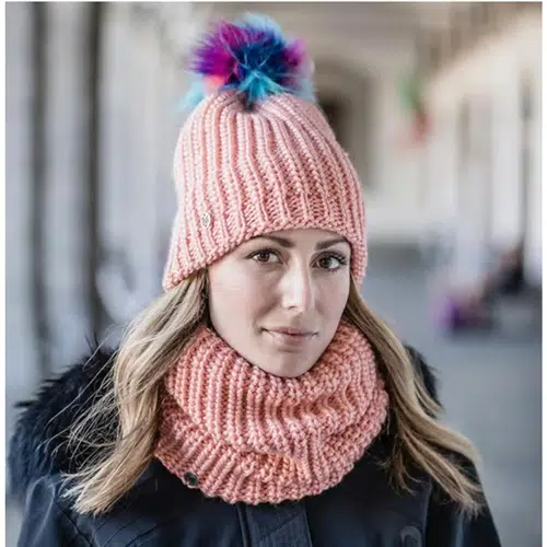 Una persona que lleva un gorro de punto rosa que recuerda al Gorro Buff Tricot y Polar Dania Peach, adornado con pompones de colores, junto con una bufanda gruesa a juego, está de pie en un pasillo al aire libre. La persona está vestida con un abrigo negro, con un fondo de elegante arquitectura arqueada.