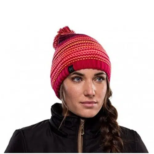 Una persona que luce un gorro colorido de punto Buff Tricot y Polar Neper Bright Pink adornado con un pompón mira hacia delante. Su pelo largo y oscuro trenzado contrasta perfectamente con la chaqueta negra con cremallera, mientras que el gorro presenta llamativos diseños en rojo y rosa.