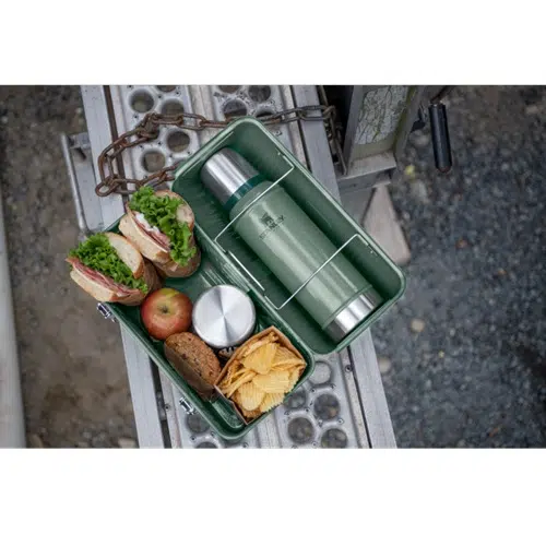 Una lonchera verde Lonchera Stanley Classic Lunch Box abierta reposa sobre una superficie de metal, mostrando su contenido: un termo, un sándwich con lechuga y pan integral, una manzana, papas fritas y un pequeño recipiente redondo. Una cadena es parcialmente visible junto a la lonchera, lo que le da un toque de encanto vintage.