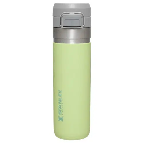 Se muestra una taza de viaje Stanley Quick Flip de 709 ml de color verde claro con tapa gris. La taza tiene un diseño elegante y cilíndrico con una tapa abatible segura y con cerradura. El logotipo y el nombre de la marca están impresos en azul en el lateral.