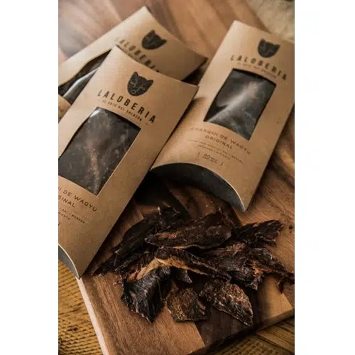 Sobre una superficie de madera se disponen los paquetes de "Charqui de Wagyu LaLobería 40grs". Al abrir el paquete se ven trozos de cecina marinada oscura que se derraman en el frente. El empaque está hecho de material ecológico de color marrón.