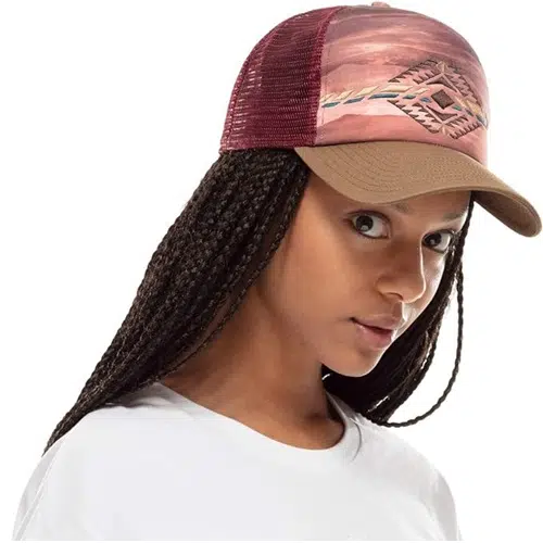 Una persona con el pelo largo trenzado lleva una gorra Jockey Buff Trucker Sykora Maroon, caracterizada por su estampado geométrico. Está vestida con una camisa blanca impecable y mira directamente a la cámara.