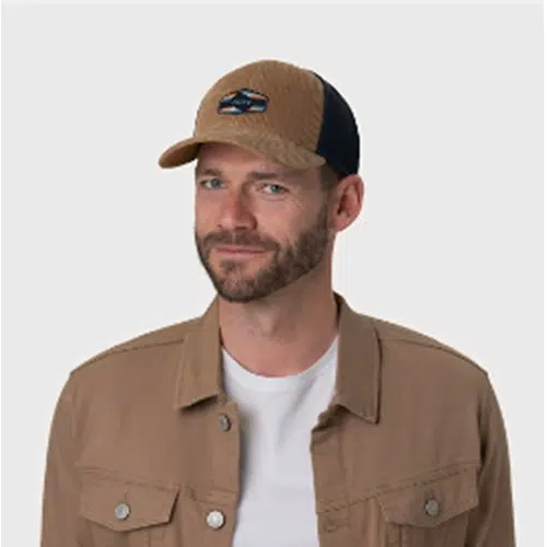 Un hombre con barba lleva una gorra de béisbol Jockey Buff Trucker Tinai Camel y una chaqueta a juego sobre una camiseta blanca. Está de pie sobre un fondo gris claro y sencillo, con un estilo natural, como un jinete preparado para la carrera.