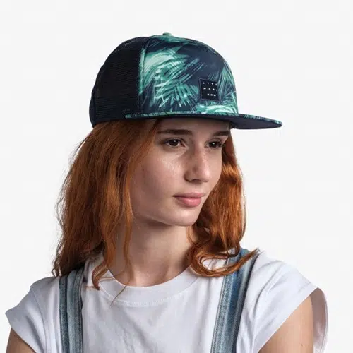 Una persona con cabello largo y rojo lleva una gorra Jockey Buff Trucker Kingara Night Bluel estampada en azul oscuro y verde combinada con una camisa blanca y un overol de mezclilla. El fondo blanco liso enfatiza los colores vibrantes de su atuendo.