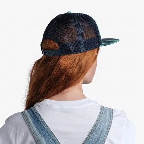 Una persona de pelo largo y rojo lleva una gorra de béisbol Jockey Buff Trucker Kingara Night Blue puesta al revés. Está vestida con una camiseta blanca y un mono de mezclilla azul y se encuentra de pie sobre un fondo blanco liso. No se le ve el rostro.